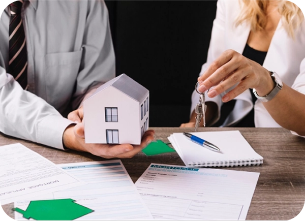 A picture shows a man holding a miniature house model while a woman signs documents at a table.